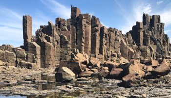 Dramatic rock formations rise against a bright blue sky, showcasing tall, angular columns. The foreground features rugged, rocky terrain.
